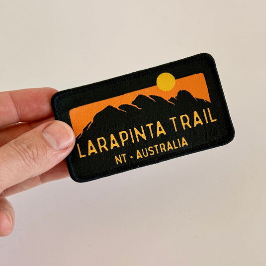 Hand holding a black iron-on patch with 'Larapinta Trail NT Australia' design on a beige background